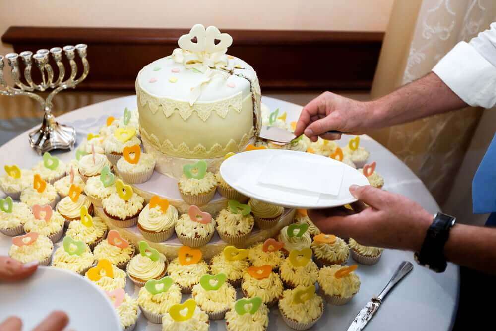 At a kosher wedding in NJ, a man is cutting a cake in front of a table full of cupcakes.