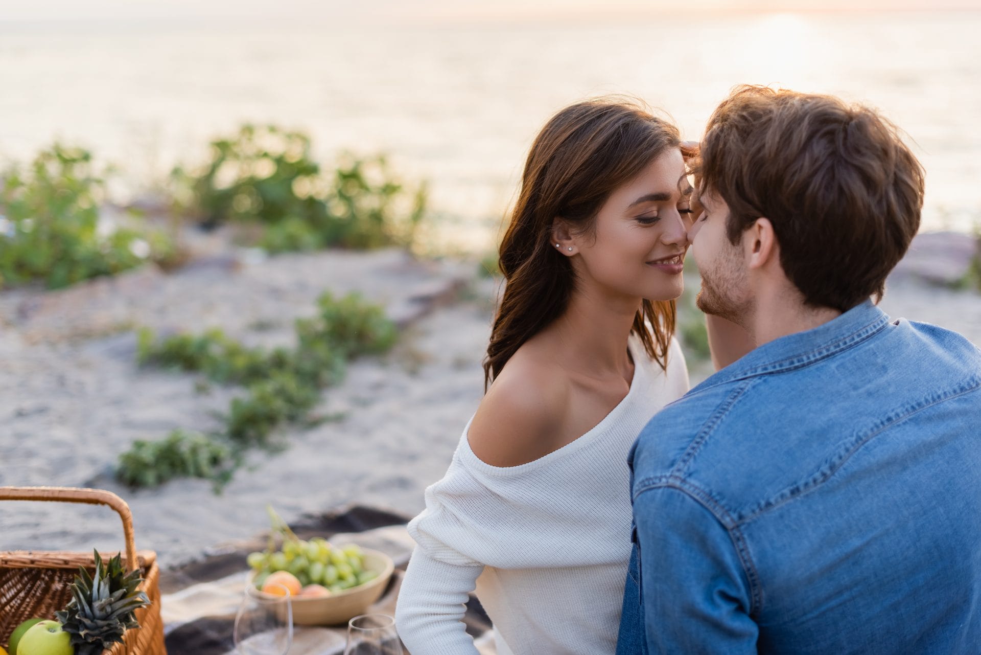 A couple enjoys a romantic moment at a beach picnic during sunset, with a picnic basket and fruits visible nearby.