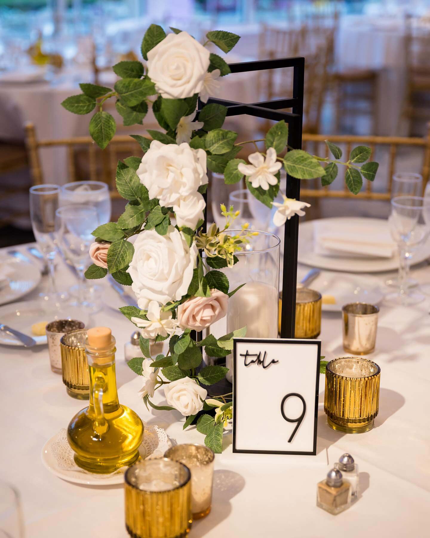table-setting-wedding-reception-at-brooklake Elegant wedding table setting featuring a floral centerpiece with a black number 9 sign, surrounded by gold candle holders and small bottles of olive oil, perfect for celebrating after getting your marriage license in NJ.