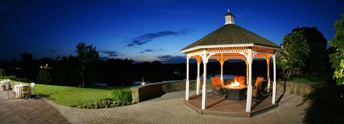 brooklake-author-page-lakeside-fire A gazebo with a fire pit inside is lit up at dusk. It's surrounded by a lawn, patio furniture, and a scenic view of trees and a pond.