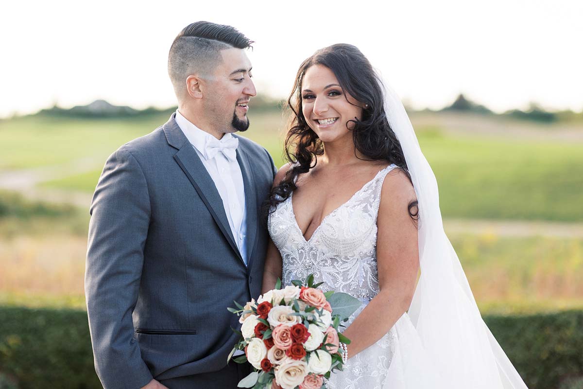 A bride and groom standing together outdoors, with the bride holding a bouquet of flowers and the groom wearing a gray suit with a white bow tie, perhaps reminiscing about the excitement of getting their marriage license in NJ.