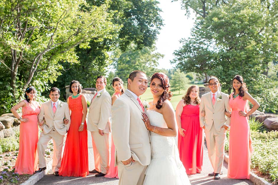 A bride and groom stand together with their bridal party in pink and beige ensembles. They are outdoors in a sunlit, green garden setting, perhaps discussing getting your marriage license in NJ amidst the joyful celebration.