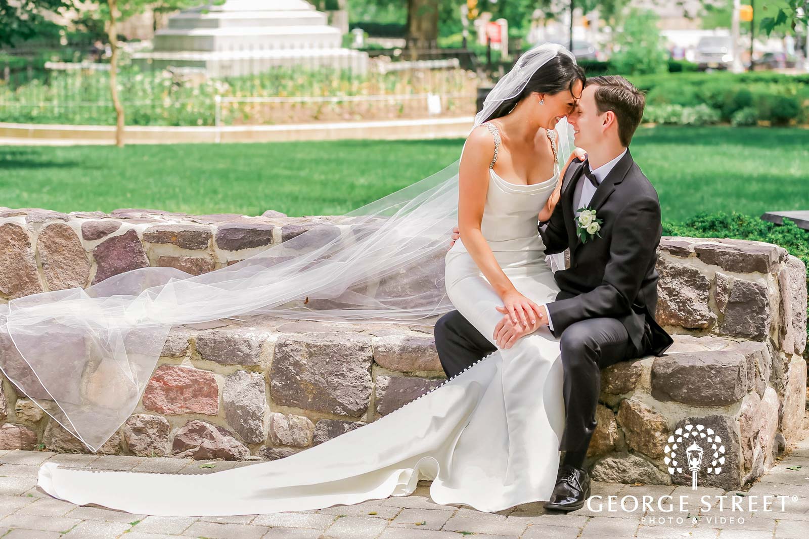 A bride and groom sit on a stone wall, with the bride's long veil flowing to the side and greenery in the background, celebrating after getting their marriage license in NJ.