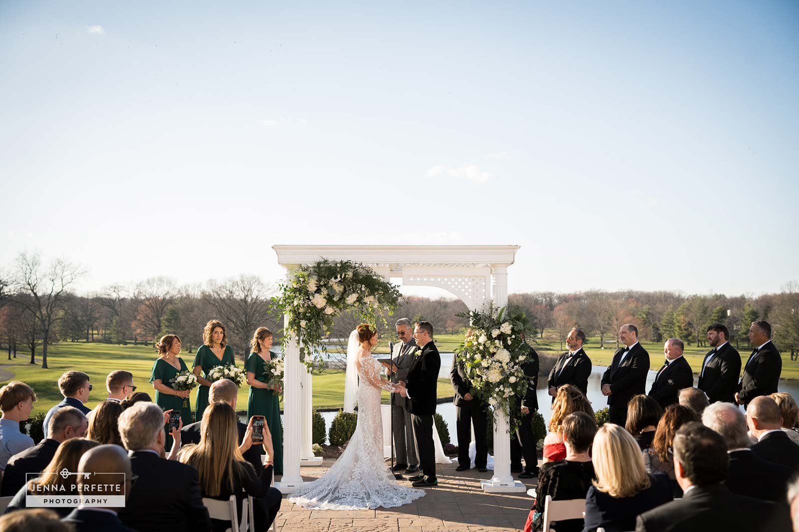 A wedding ceremony takes place outside under a decorated white arbor, with the couple—who recently completed getting their marriage license in NJ —exchanging vows in front of their bridal party and seated guests on a sunny day.