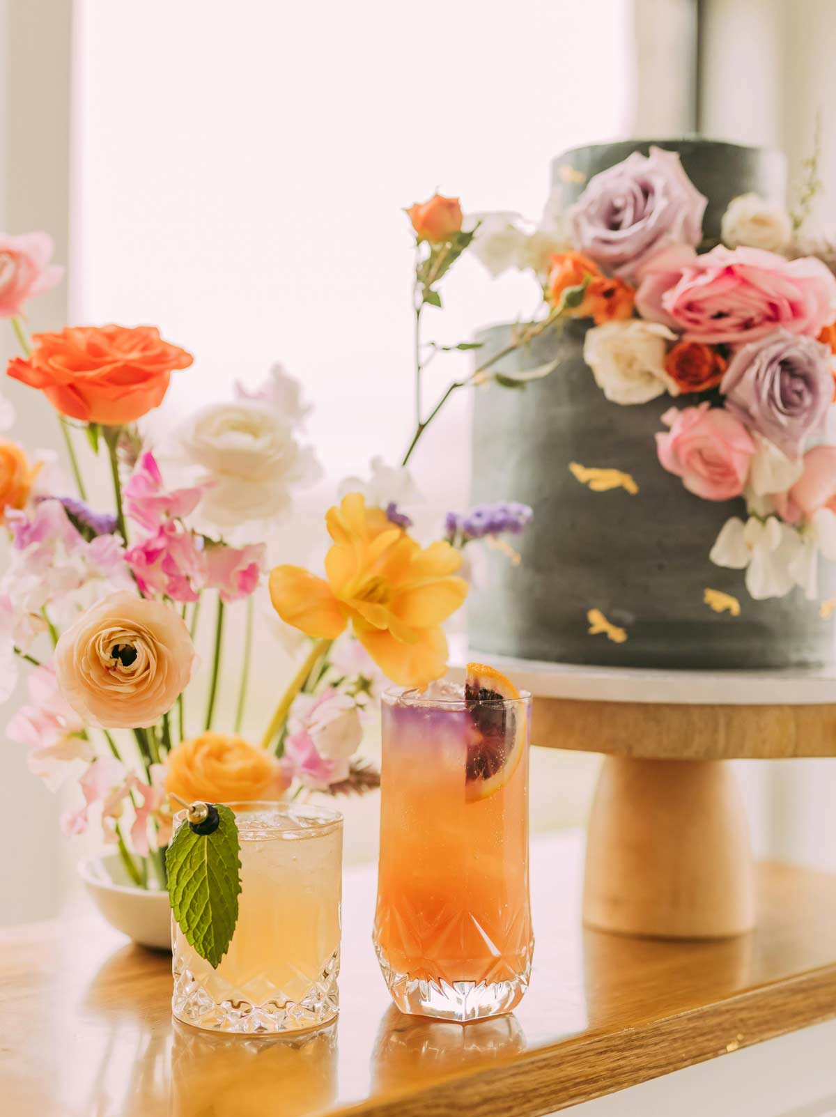Two colorful cocktails sit on a table adorned with fresh flowers, beside a dark frosted cake decorated with pastel flowers on a stand, perfect for celebrating moments like getting your marriage license in NJ.