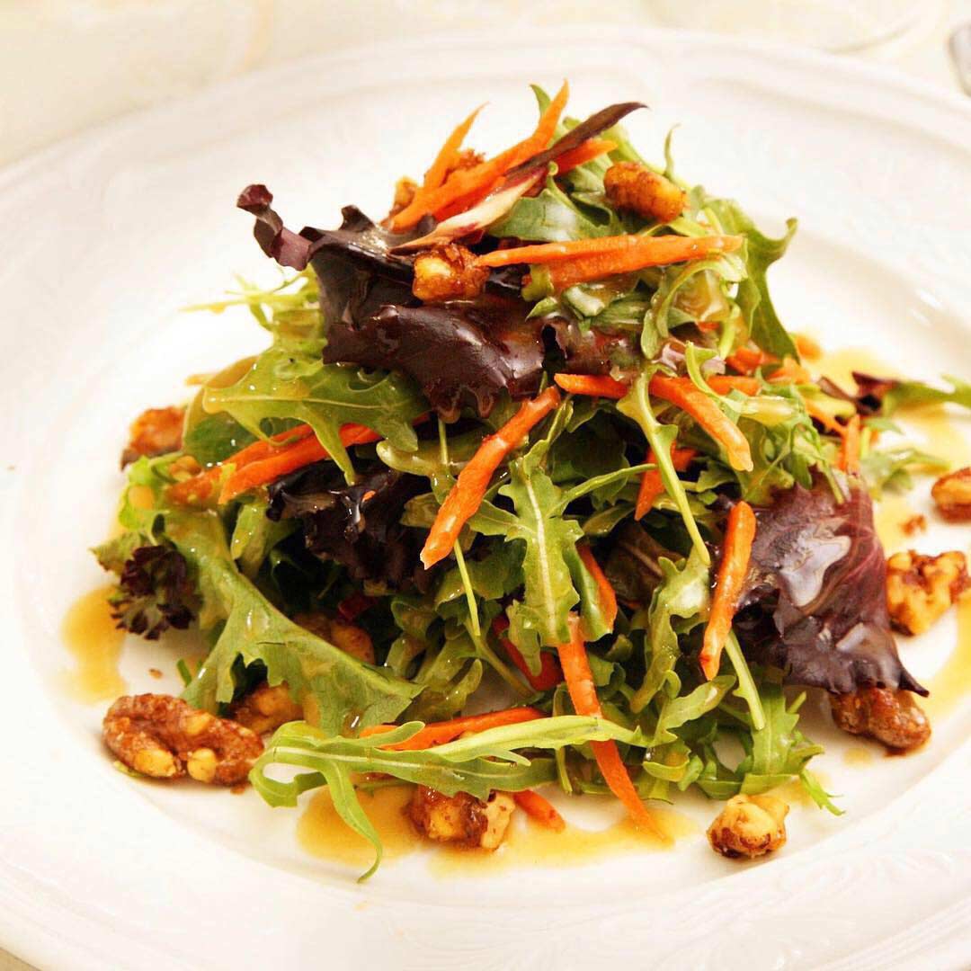 A mixed greens salad with grated carrots and candied nuts on a white plate, drizzled with dressing, epitomizes the refined Cuisine at Brooklake.