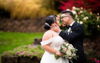 A bride and groom, dressed in a white gown and black suit respectively, share a tender moment outdoors, surrounded by the lush greenery and vibrant flowers of Cuisine at Brooklake. The groom holds the bride's face gently.