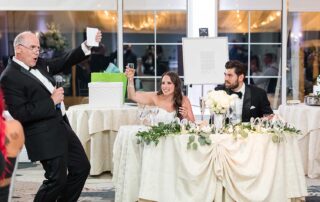 At Love Stories at Brooklake, a man in a tuxedo makes a toast with a microphone as the seated bride and groom raise their glasses, surrounded by elegantly decorated tables.