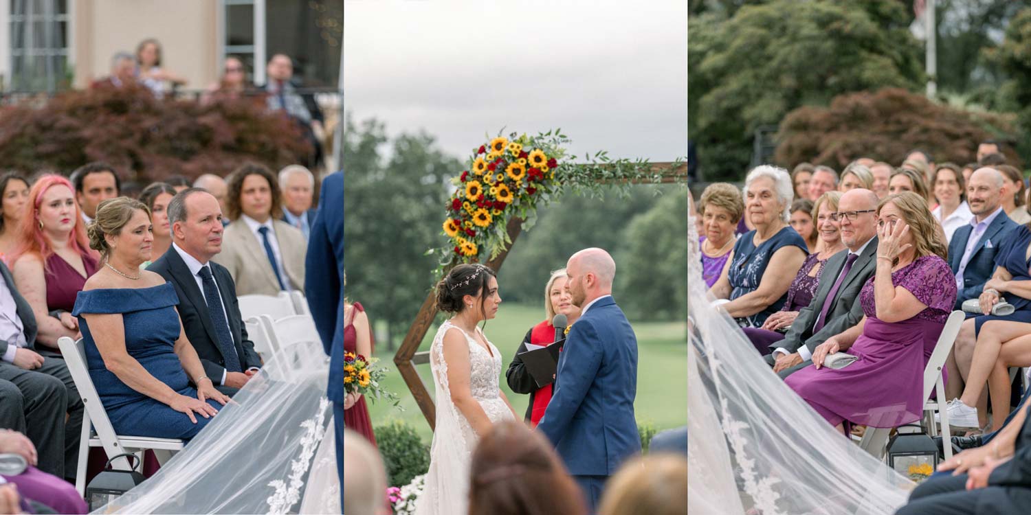 A couple is getting married outdoors at Love Stories at Brooklake. Guests are seated on each side, while a floral arch frames the scene. A woman in a red outfit officiates the ceremony with elegance and warmth, making it an unforgettable celebration of love.
