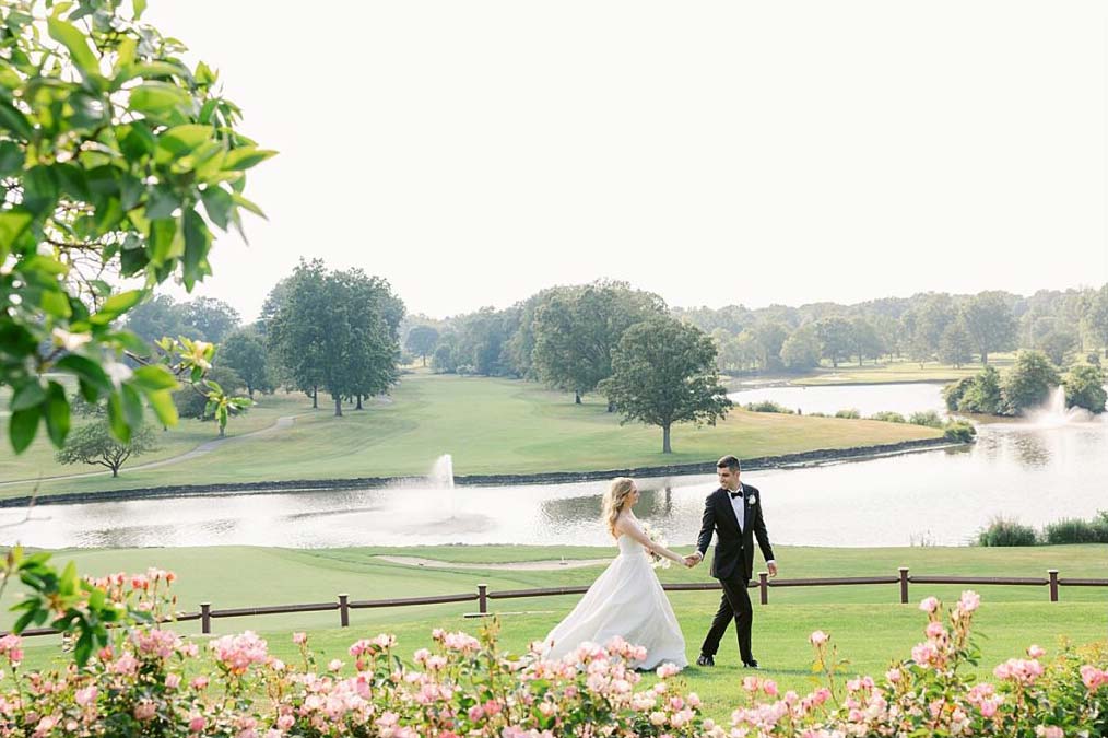 A bride and groom walk hand in hand across a lush green lawn, honoring parents and grandparents at their wedding. With a lake and trees in the background, pink flowers bloom in the foreground, adding to the moment's beauty.