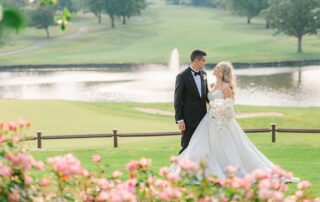 A couple in formal attire stands on a grassy area, honoring parents and grandparents with a fountain and trees in the background. Pink flowers bloom gracefully in the foreground.
