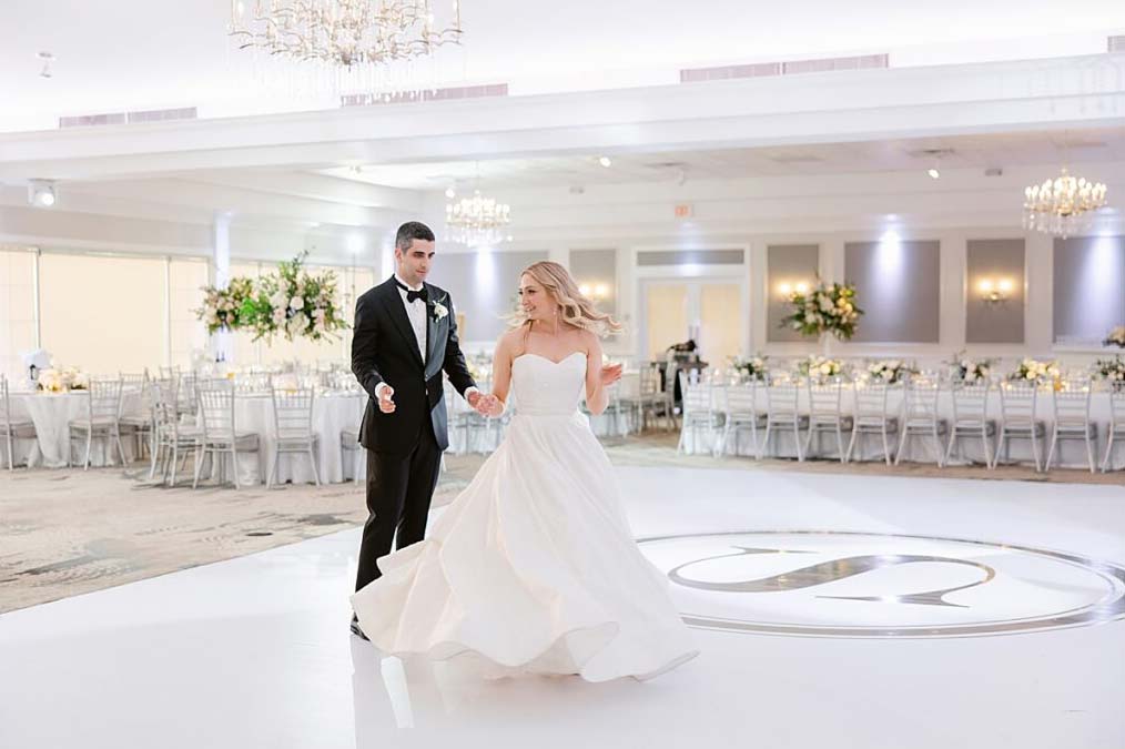 A couple gracefully dances on a white floor in a large, elegantly decorated room with a chandelier and tables set for an event, honoring parents and grandparents at their wedding with each step they take.