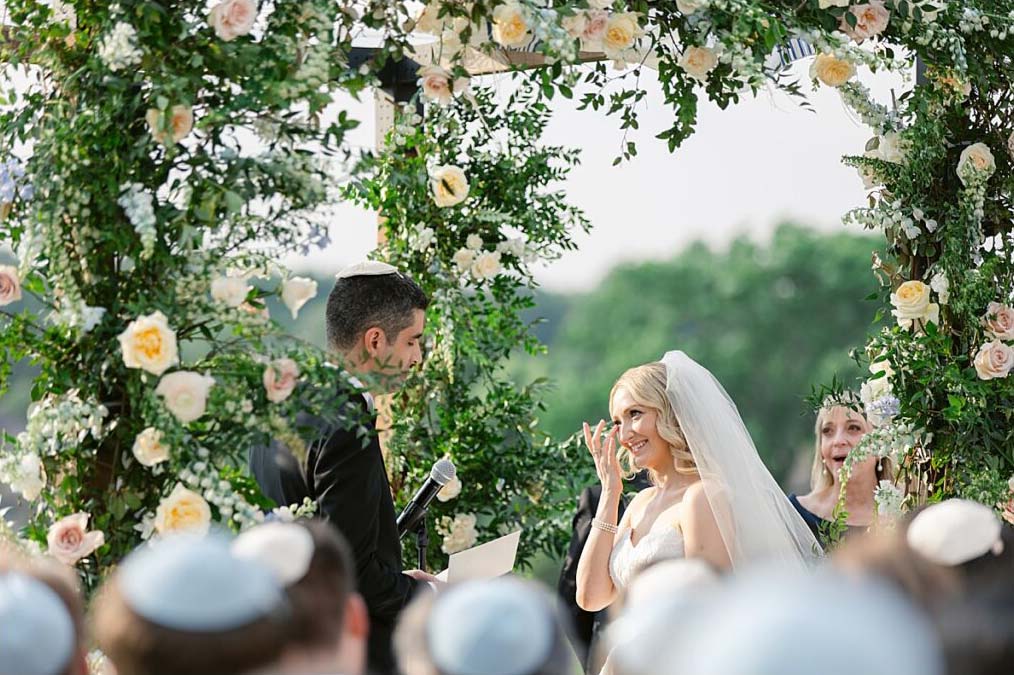 laurenkearns-brooklake-bride-groom-wedding-ceremony-0053 A bride and groom stand under a floral arch during an outdoor wedding ceremony, with the bride wiping her eye. Guests wearing yarmulkes are visible in the foreground, reflecting on the couple honoring parents and grandparents at their wedding.