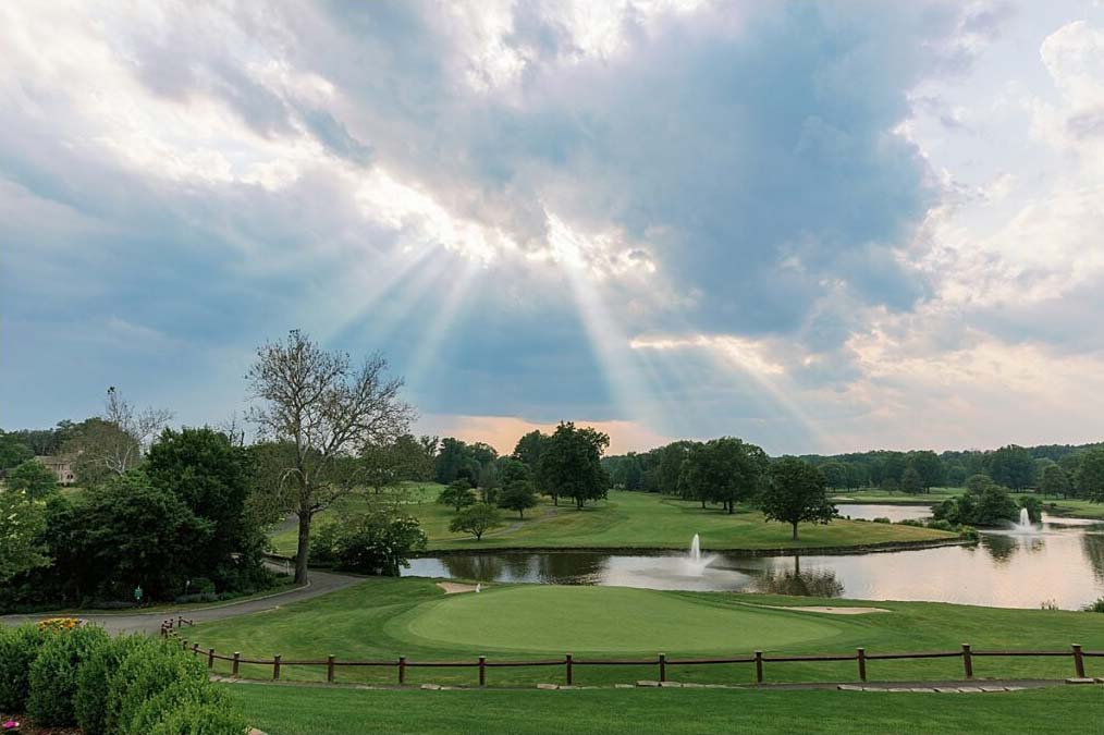 The golf course landscape, perfect for honoring parents and grandparents at your wedding, features a lush green with trees and a serene pond. Sun rays break through the clouds, casting a warm light onto this cherished setting.