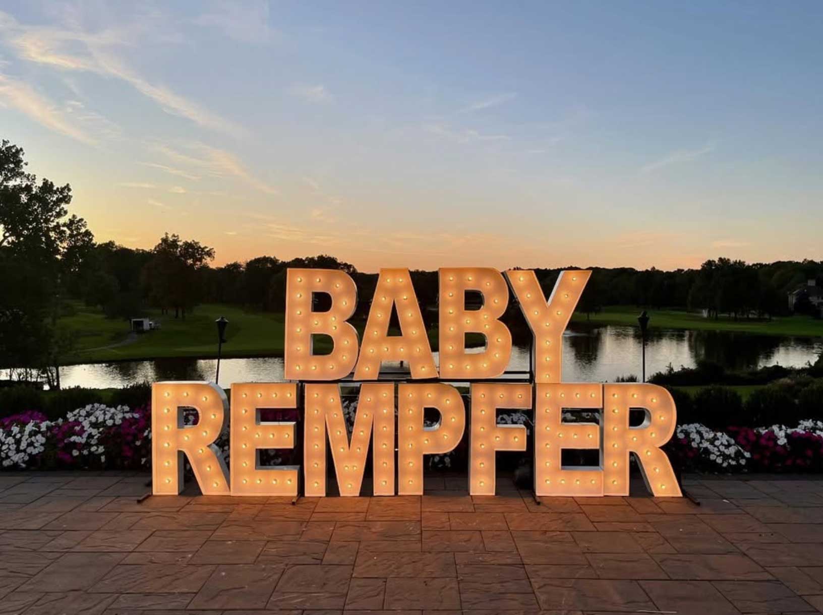 lighted-baby-side-lakeside-brooklake-shower Illuminated marquee letters spell "BABY REMPFER" on a serene patio, capturing the essence of NJ Milestone events against a backdrop of a shimmering lake and vibrant sunset.