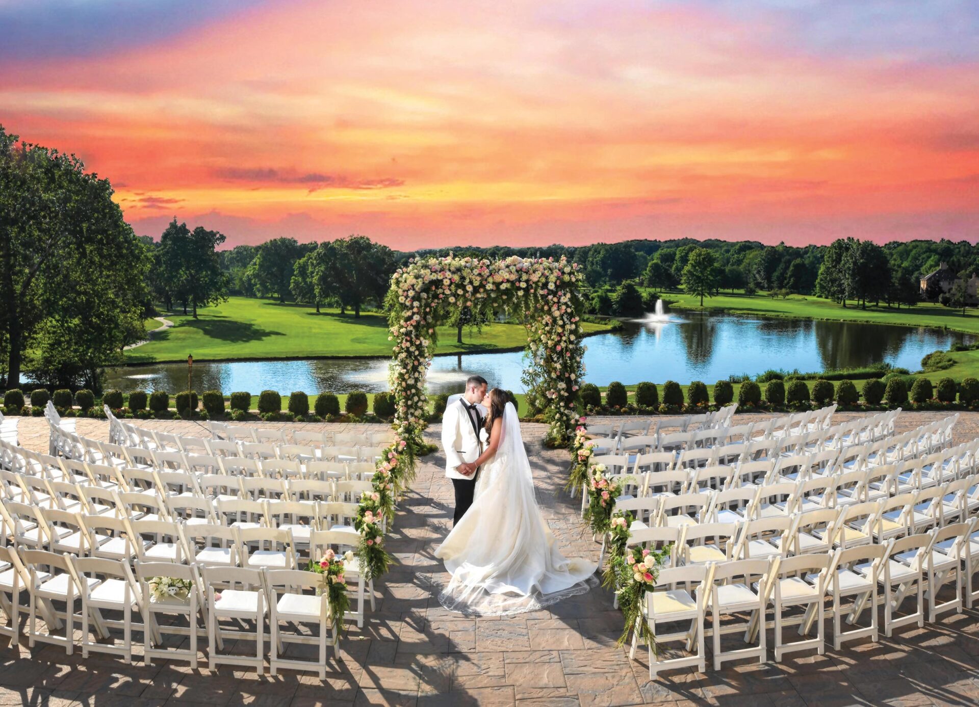 Bride and groom kiss under a floral arch at a stunning NJ wedding venue. Rows of white chairs line the pathway, while a pond and sunset sky provide the perfect backdrop.