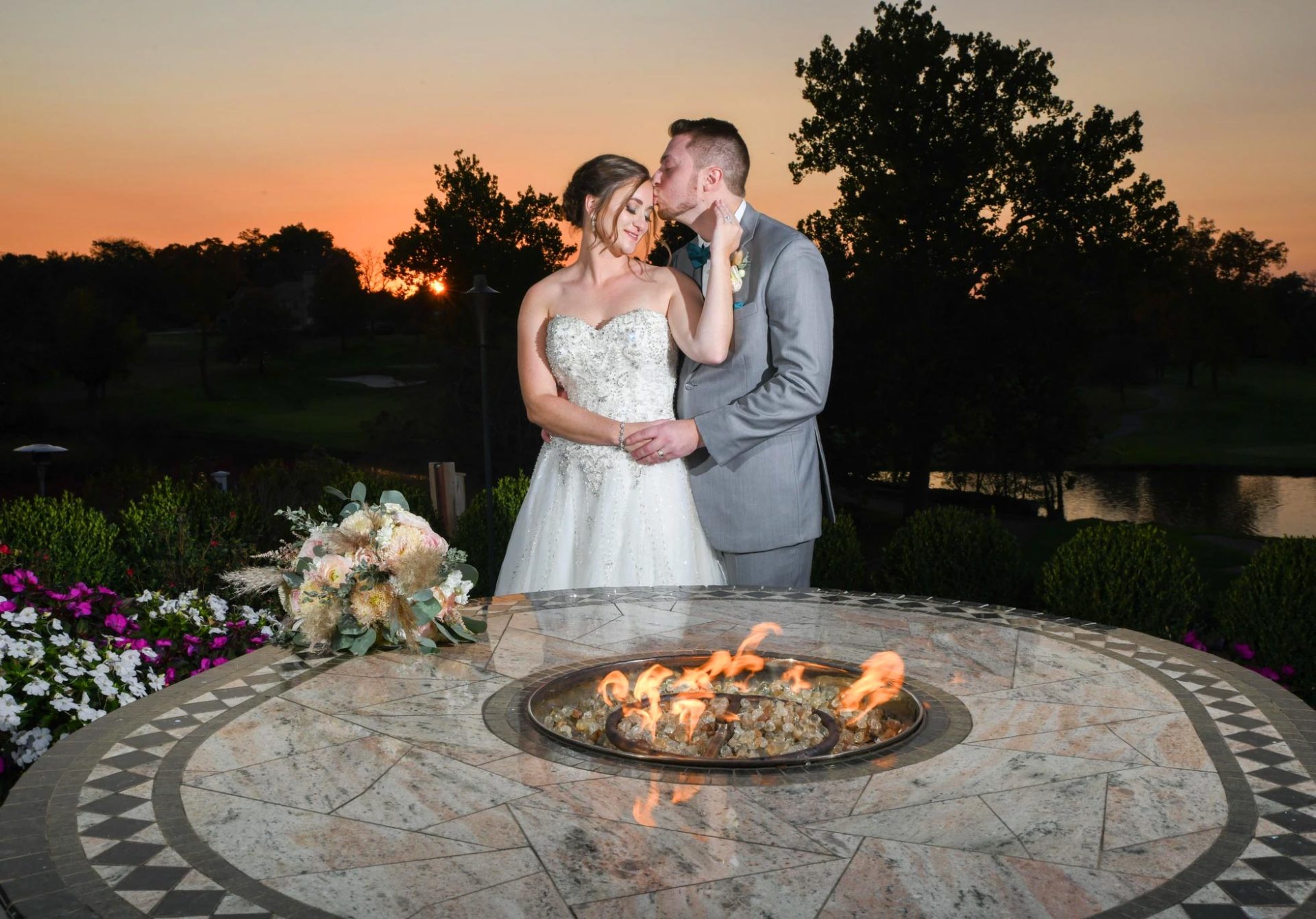 bride-and-groom-at-brooklake-firepit A couple in wedding attire poses by a circular fire pit at sunset at their beautiful NJ wedding venue, with the groom gently kissing the bride's forehead. A bouquet and vibrant flowers are visible in the foreground, adding an extra touch of romance to the scene.