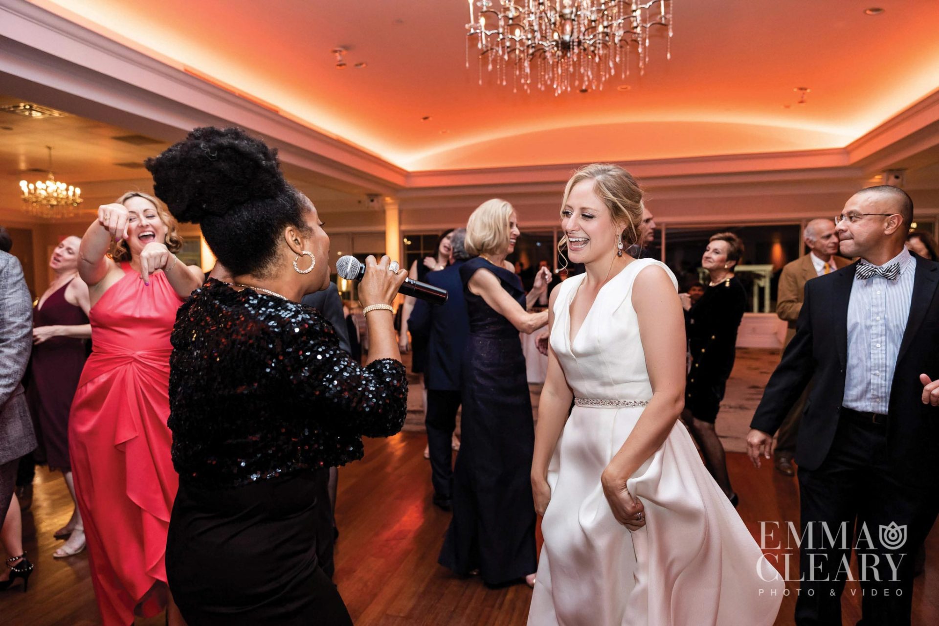singer-and-bride-on-brooklake-dance-floor A woman in a white dress stands and smiles as another woman sings into a microphone on the dance floor at an elegant NJ wedding venue. Other attendees stand and converse in the background, adding to the festive atmosphere.