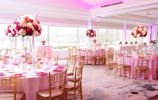 A Vibrant & Unforgettable Bar Mitzvah Celebration at Brooklake: Ayla’s Big Day featured a decorated banquet hall with round tables covered in pink linens, gold chairs, and large floral centerpieces in shades of white, pink, and red.