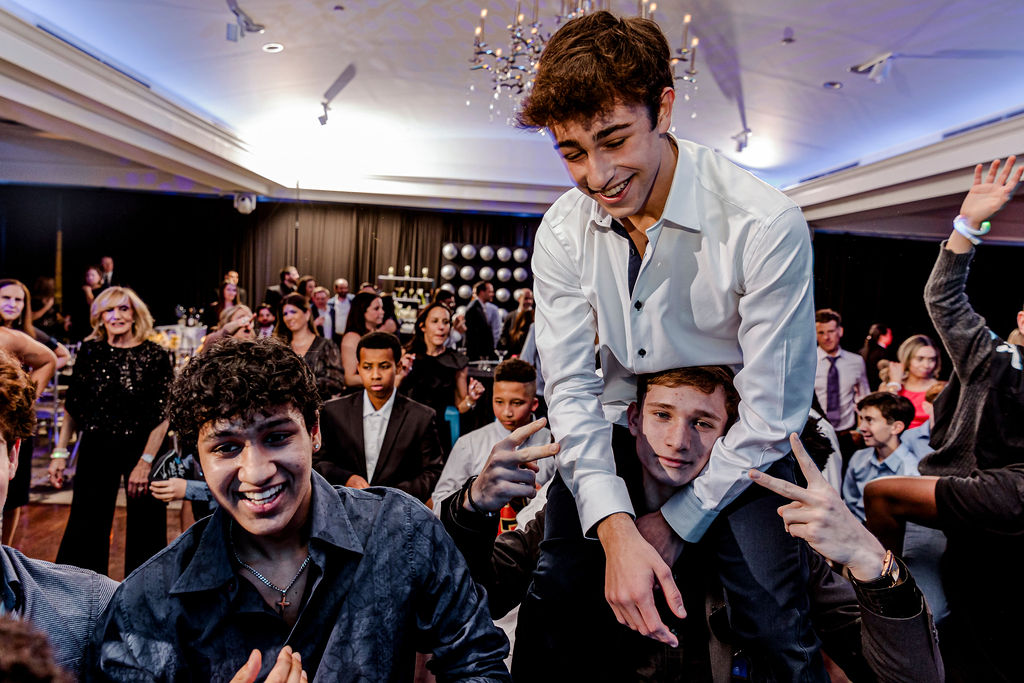 MasonRheingoldBarMitzvahReception-NewJerseyBarMitzvahPhotography-JoeCurryPhotography-2025-1144 A group of teenagers at a party in an NJ wedding venue; one boy sits on another's shoulders while others smile and flash peace signs, with more people and elegant decorations in the background.