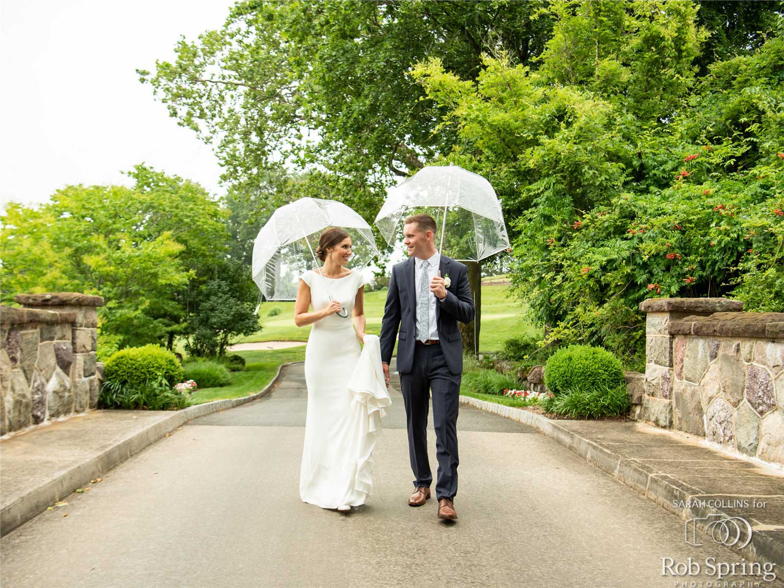 bride-and-groom-on-brooklake-bridge with umbrellas A bride and groom walk together on a paved path at an NJ wedding venue, holding clear umbrellas, surrounded by greenery and stone walls on a cloudy day.
