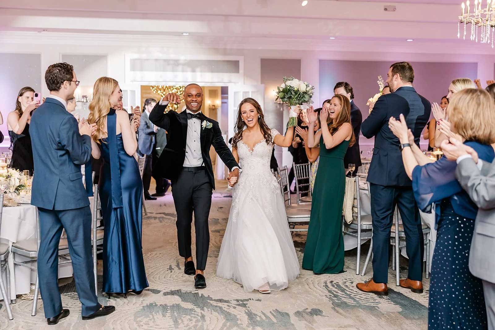 bride-groom-entering-wedding-reception-brooklake A bride and groom walk hand in hand into an elegant NJ wedding venue as guests stand, clap, and cheer around them.