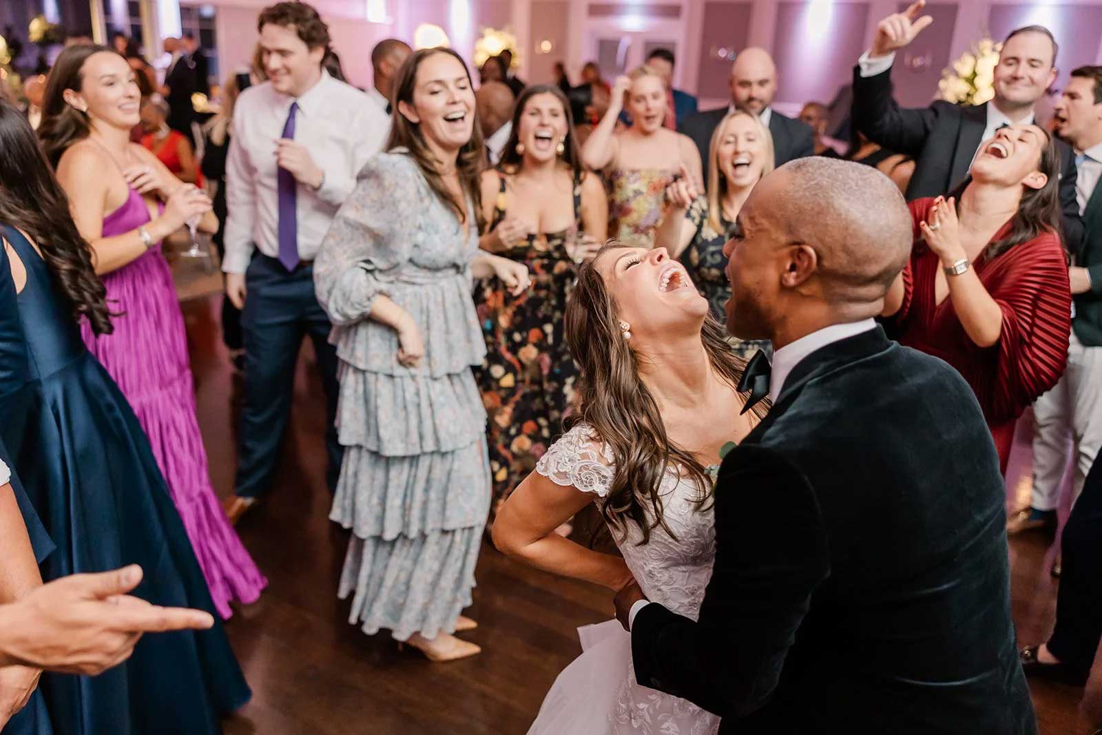bride-groom-guests-dancing-wedding-reception-brooklake A bride and groom laugh and dance together on a crowded dance floor surrounded by cheering, smiling guests at a beautiful NJ wedding venue.