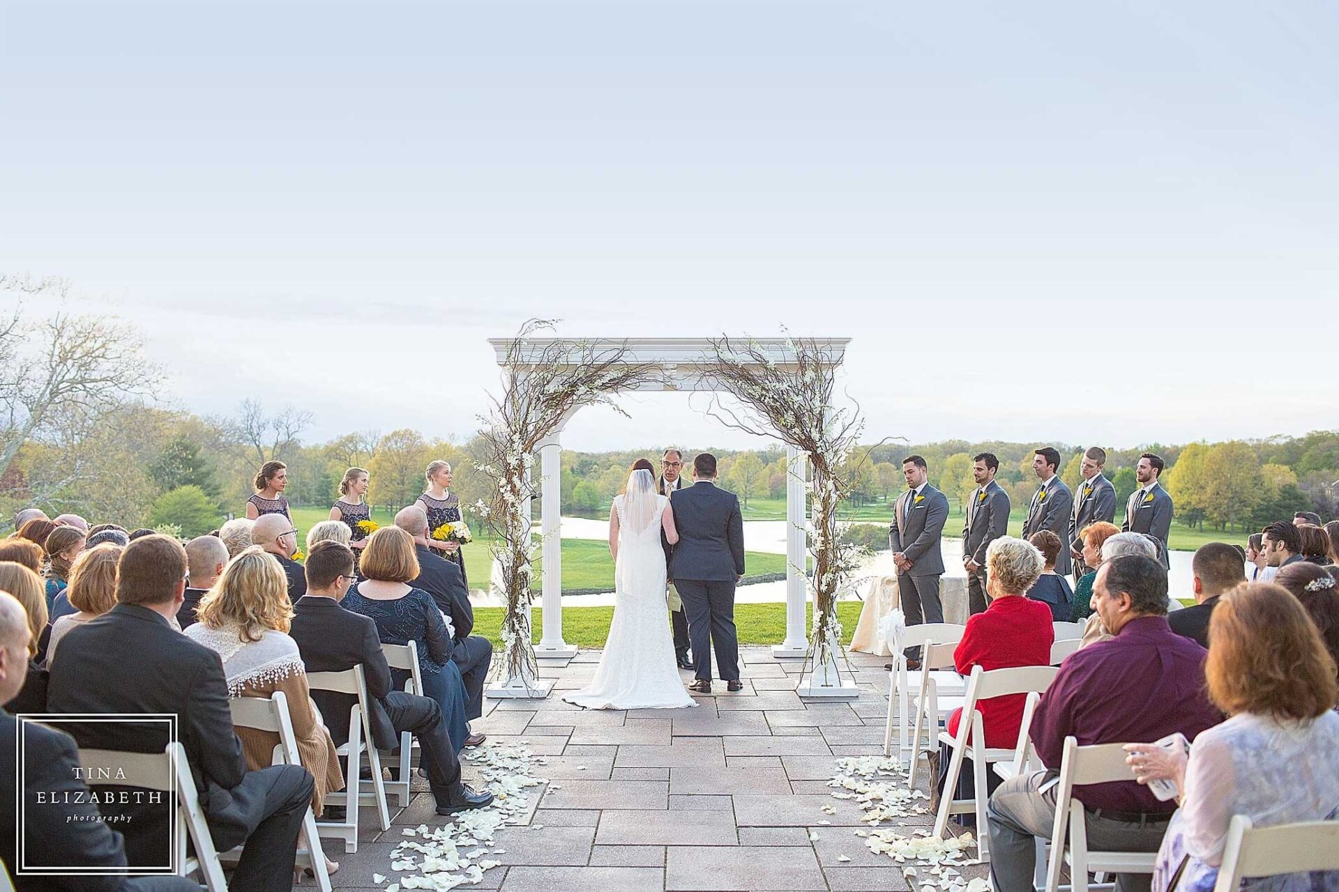 fall-outdoor-wedding-ceremony-brooklake A couple stands under a decorated arch at a stunning NJ wedding venue during an outdoor ceremony, surrounded by their wedding party and seated guests on a paved patio with a scenic landscape in the background.
