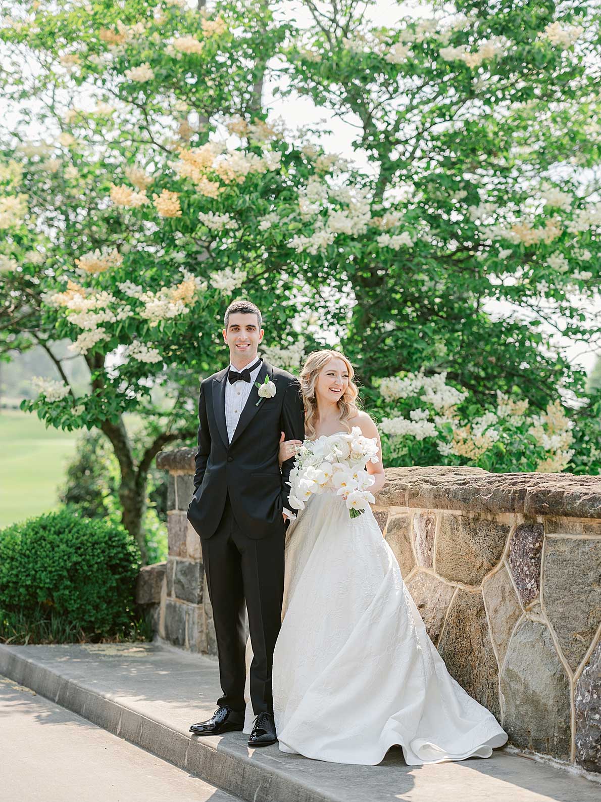 lauren-kearns-bride-groom-on-bridge-at-brooklake A bride and groom stand side by side outdoors near a stone wall at an elegant NJ wedding & event venue, dressed in formal wedding attire, with greenery and blooming trees in the background.