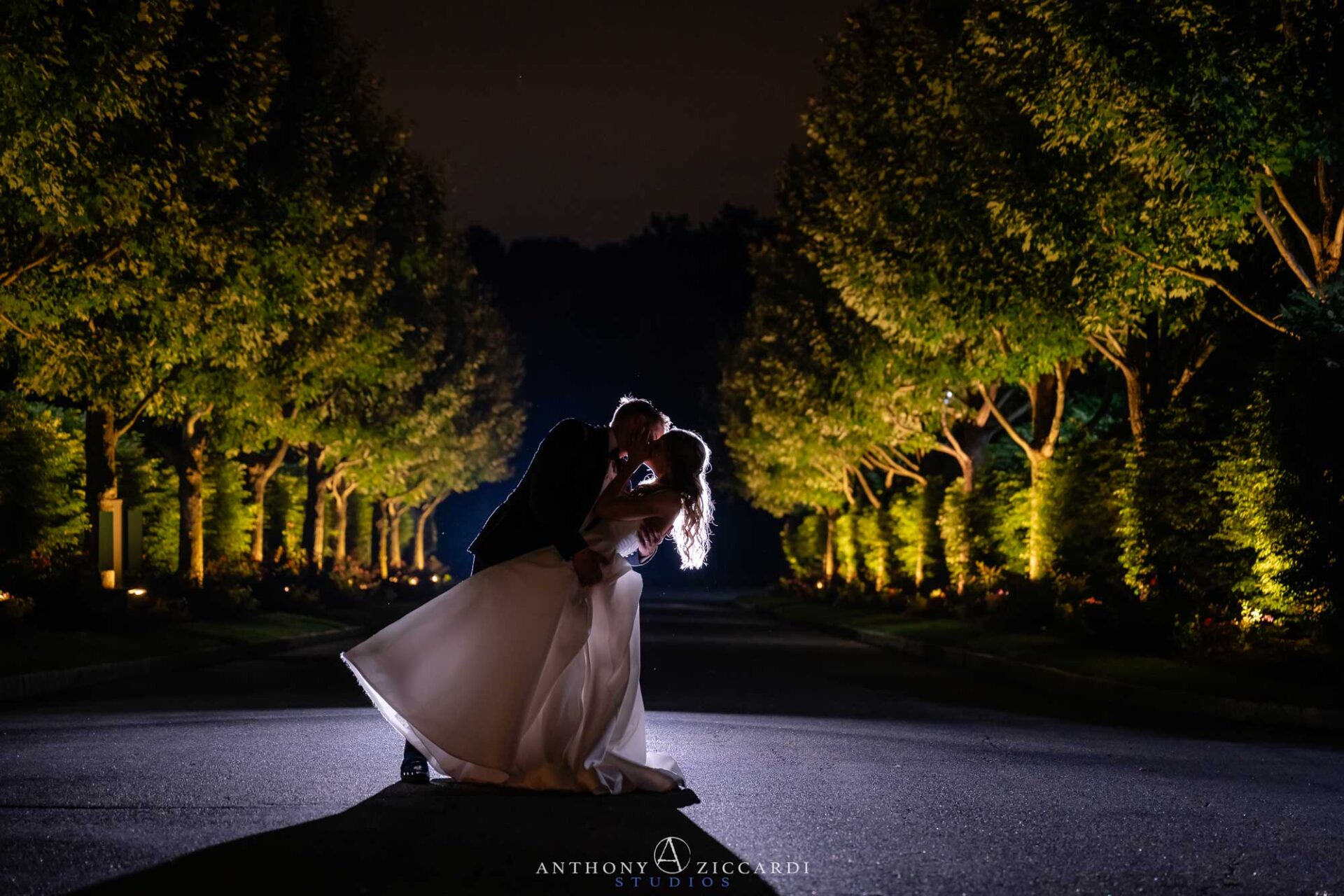 nighttime-backlit-bride-and-groom-brooklake A bride and groom share a kiss at night on a tree-lined road outside their NJ wedding venue, illuminated from behind by bright lights, casting long shadows on the pavement.