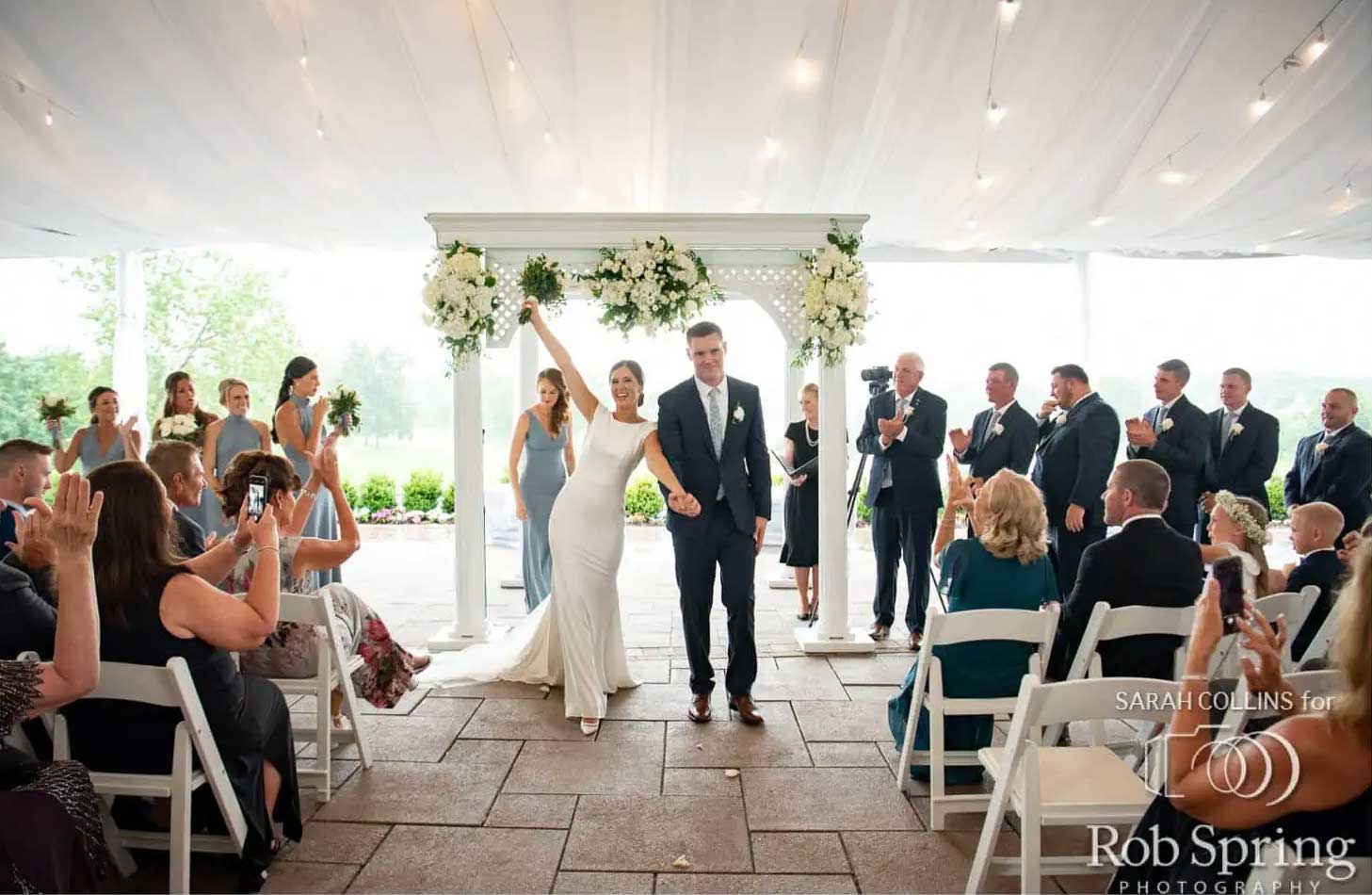 patio-covered-wedding-ceremony-brooklake A bride and groom walk down the aisle at their NJ wedding venue, smiling and holding hands, surrounded by applauding guests seated on both sides.