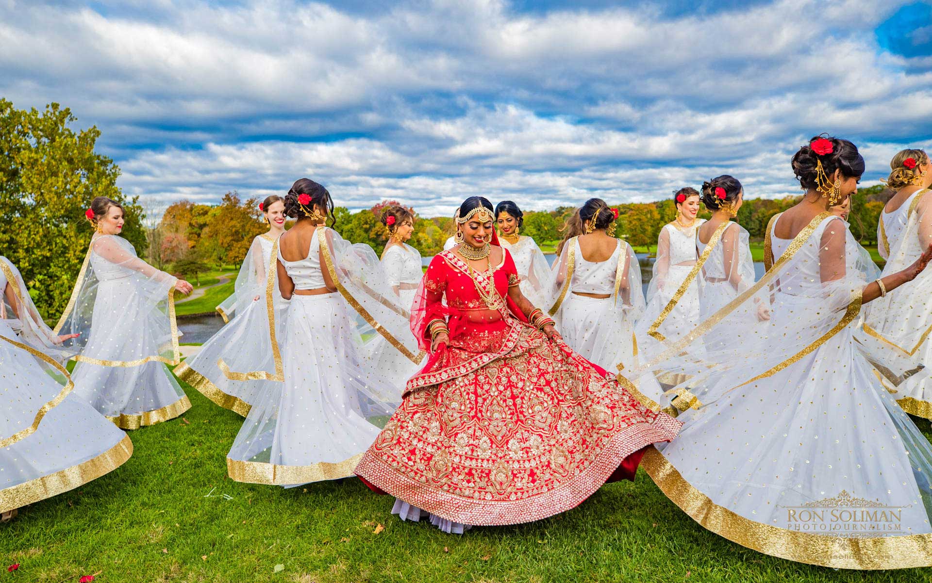 BROOKLAKE-COUNTRY-CLUB-INDIAN-WEDDING-11 A bride in a red and gold lehenga stands in the center while women in white and gold dresses dance around her on a grassy field under a cloudy sky, capturing the vibrant spirit of NJ Teen Events.