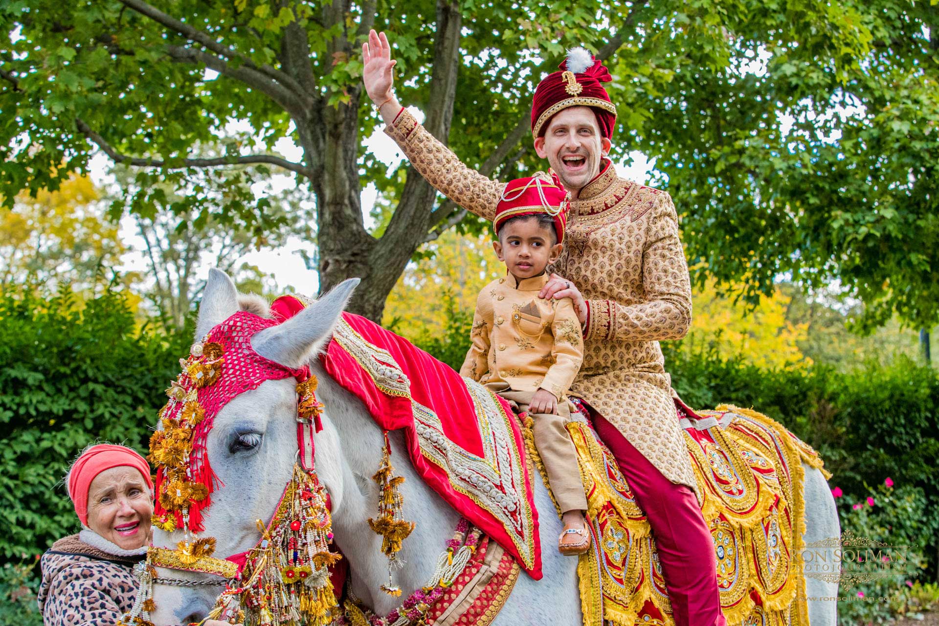 BROOKLAKE-COUNTRY-CLUB-INDIAN-WEDDING-18 A man and a child in traditional attire sit on a decorated white horse, waving, while a woman stands nearby. The festive scene, surrounded by green trees, captures the joy often seen at NJ Teen Events.
