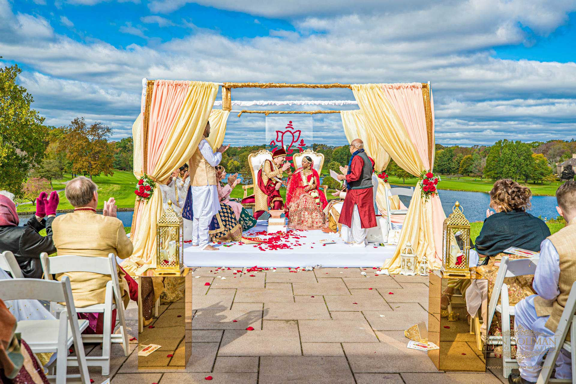 BROOKLAKE-COUNTRY-CLUB-INDIAN-WEDDING-35 A couple in traditional attire sits under a decorated canopy during an outdoor wedding ceremony, surrounded by guests and vibrant scenery—an elegant celebration reminiscent of festive NJ Teen Events.