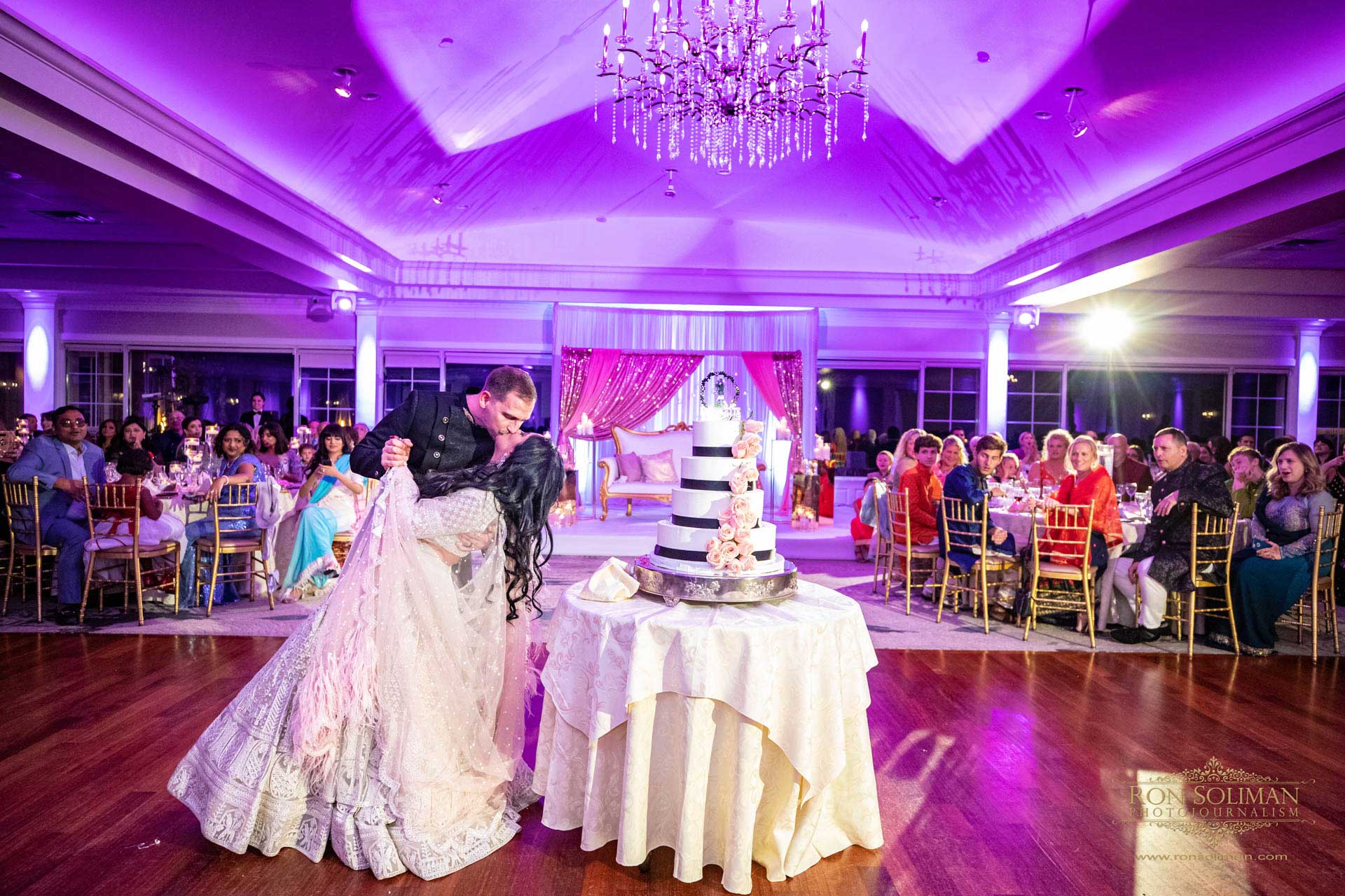 BROOKLAKE-COUNTRY-CLUB-INDIAN-WEDDING-47 A couple shares a dip on the dance floor at a wedding reception, with guests seated around and a multi-tiered cake in the foreground—capturing a magical moment reminiscent of joyful NJ Teen Events.