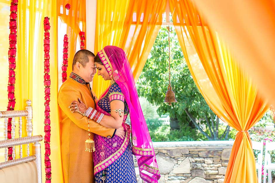 Brooklake-Country-Club_0038 A couple in traditional Indian attire embraces under vibrant yellow and orange drapes, with garlands and outdoor greenery in the background—perfect inspiration for NJ Teen Events celebrating culture and style.