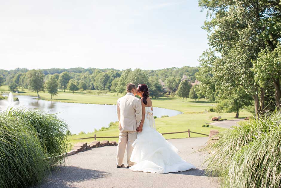 Brooklake-Country-Club_0058 A bride and groom stand together outdoors on a paved path, overlooking a pond and green landscape on a sunny day, capturing a moment as magical as NJ Teen Events.