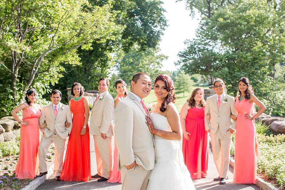 Brooklake-Country-Club_0064 A bride and groom pose outdoors with their wedding party, dressed in light suits and coral dresses, surrounded by greenery on a sunny day—capturing memories as bright as any NJ Teen Events celebration.