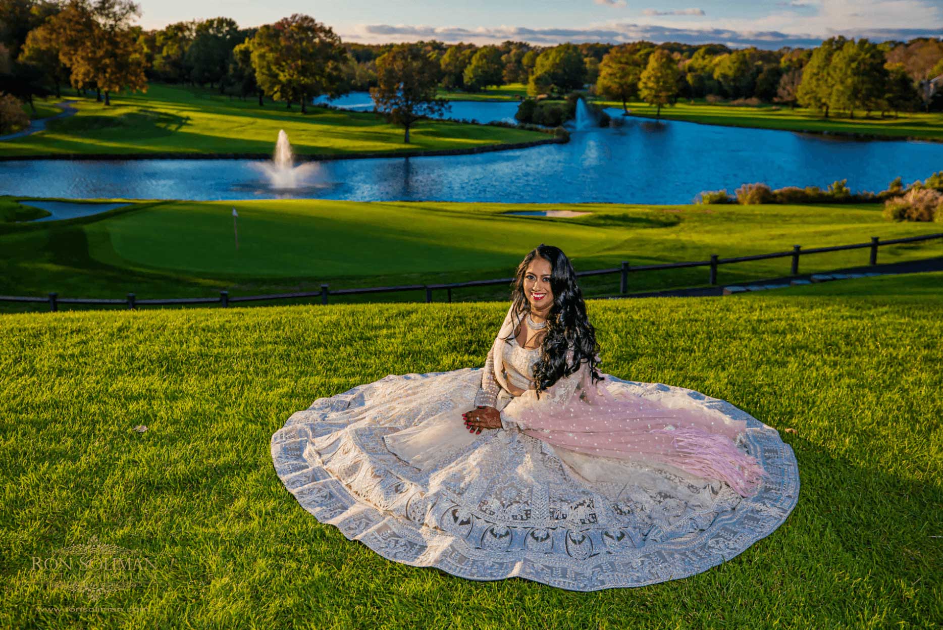 Brooklake-Indian-Wedding-Bride-on-grass A woman in a white and pink dress sits on green grass with a large pond, fountain, and trees in the background under a blue sky, capturing the serene vibe of NJ Teen Events.