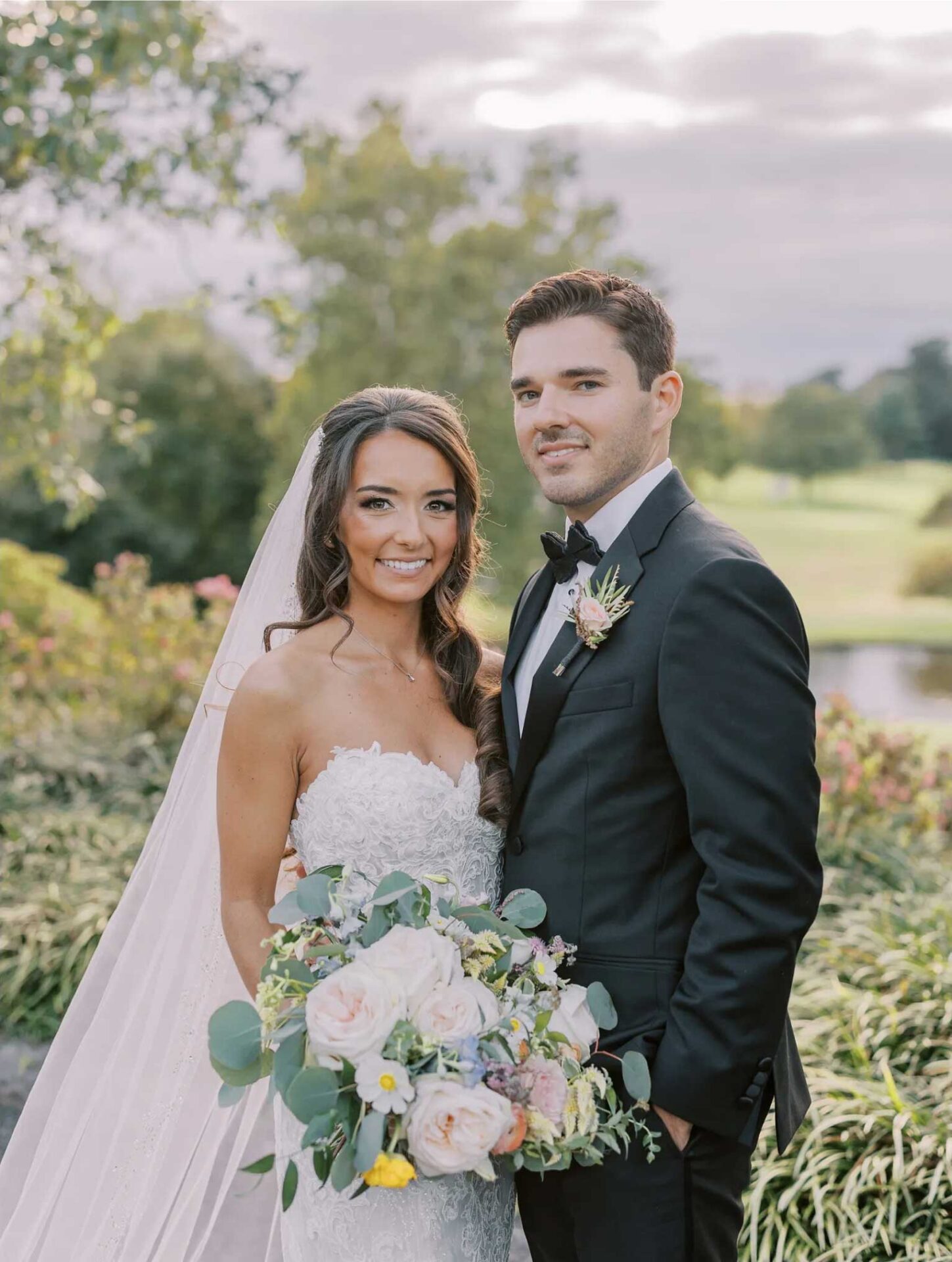 bride-groom-ready-for-photos-at-brooklake A bride in a white gown and a groom in a black suit pose together outdoors at an elegant NJ wedding & event venue, holding a bouquet of flowers, with greenery and a cloudy sky in the background.