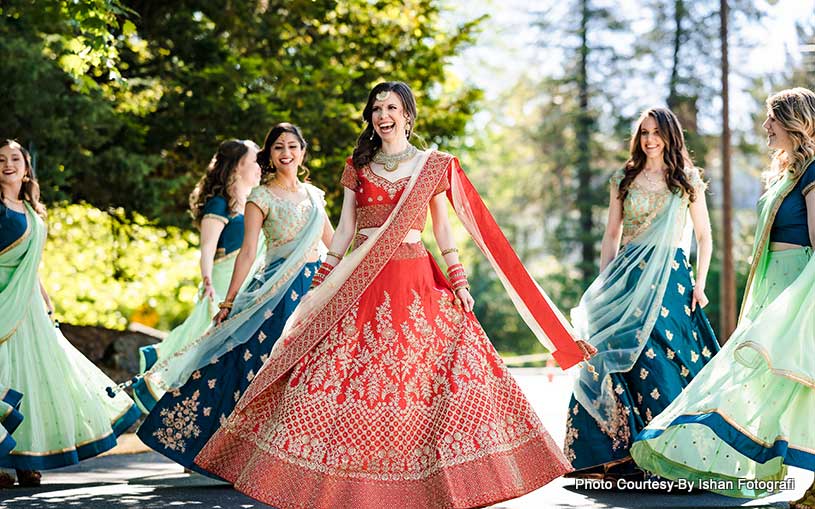 brooklake-Christine_Shivam-4 A bride in a red and gold lehenga smiles and spins at an NJ Teen Events celebration, surrounded by bridesmaids in matching teal and blue lehengas outdoors on a sunny day.