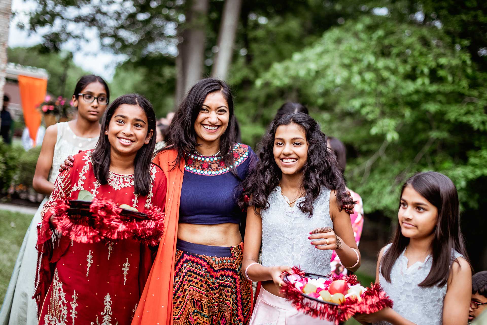 brooklake-Indian-Wedding-photographer-003 A group of smiling girls and a woman stand outdoors at an NJ Teen Events gathering, some holding trays with red garlands and items, surrounded by greenery.