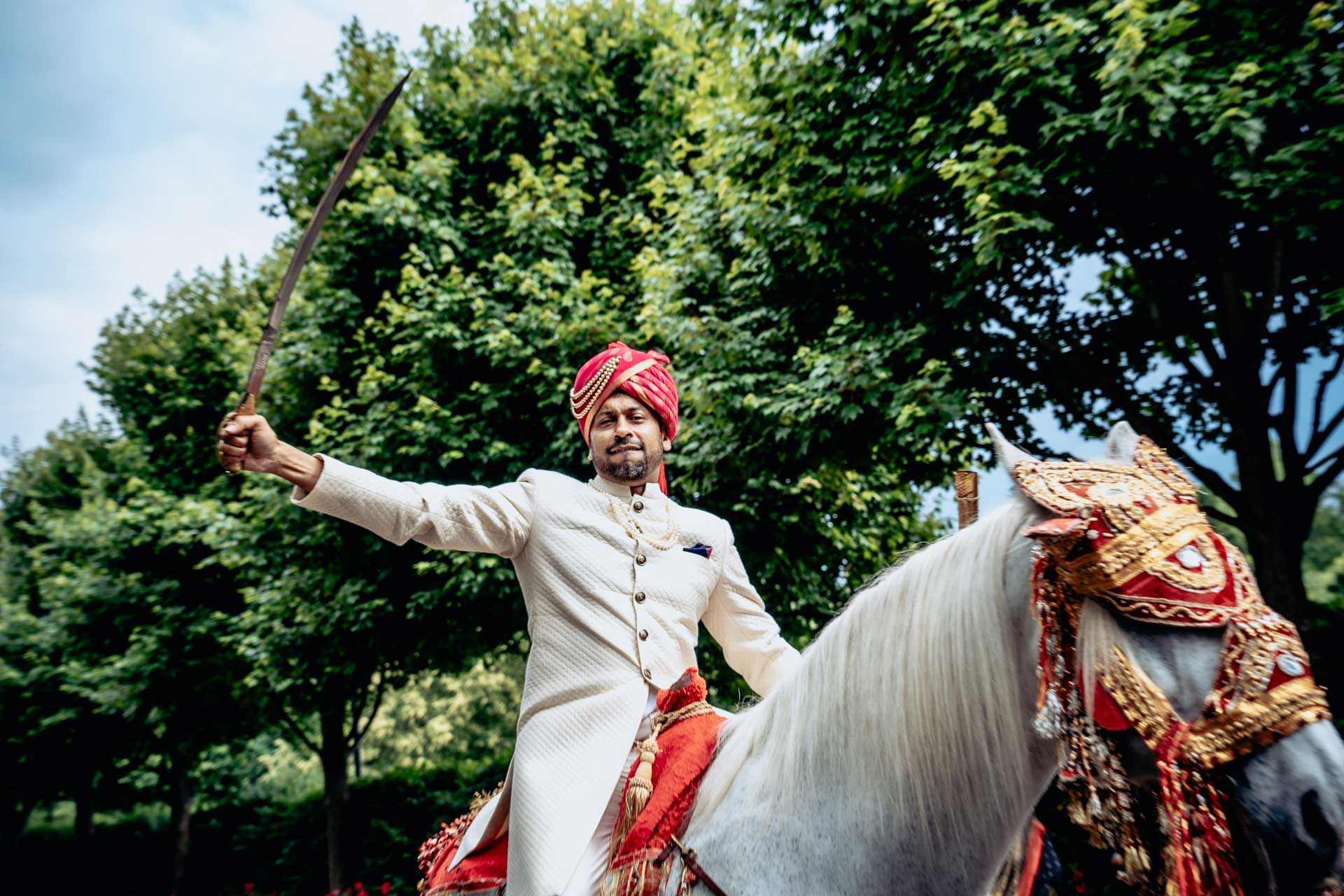 brooklake-Indian-Wedding-photographer-020 A man in a white traditional outfit and red turban rides a decorated white horse, holding a sword aloft, with green trees in the background—an inspiring scene reminiscent of vibrant NJ Teen Events.