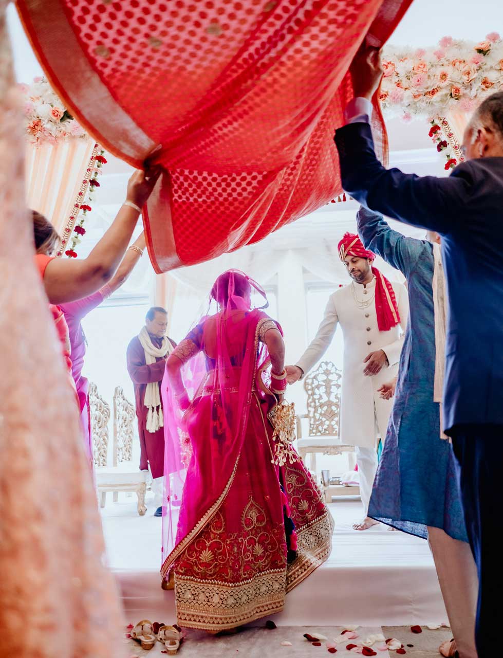 brooklake-Indian-Wedding-photographer-029 A bride in a pink saree walks towards a groom in white traditional attire during a wedding ceremony, with family members holding a red cloth above her at one of the vibrant NJ Teen Events.