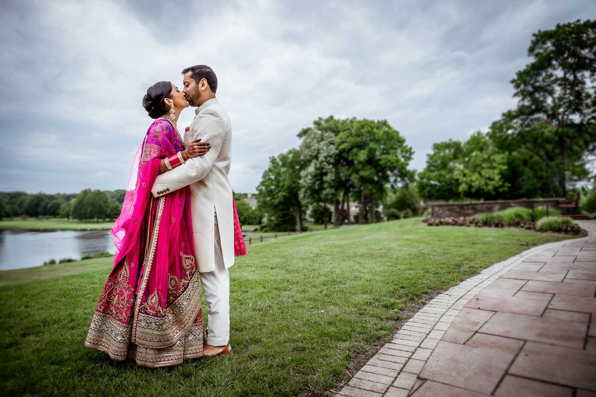 brooklake-Indian-Wedding-photographer-040 A couple dressed in traditional South Asian attire embrace and kiss outdoors on a grassy lawn near a paved walkway, capturing the joyful spirit often seen at NJ Teen Events, with trees and a cloudy sky in the background.