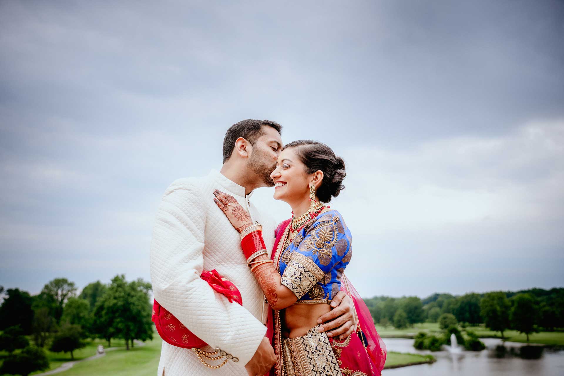brooklake-Indian-Wedding-photographer-044 A man in a white sherwani kisses a woman in a blue and gold saree on the forehead outdoors, with trees and water in the background—capturing a tender moment perfect for NJ Teen Events inspiration.