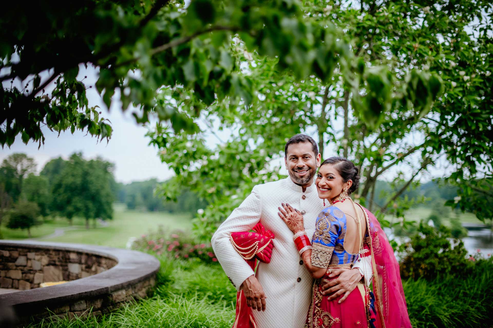 brooklake-Indian-Wedding-photographer-046 A couple in traditional South Asian attire poses outdoors near a stone wall and green trees, smiling at the camera during one of the popular NJ Teen Events.