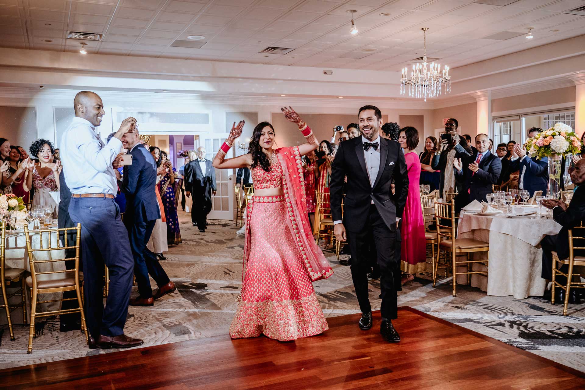 brooklake-Indian-Wedding-photographer-054-2 A couple in formal attire makes a joyful entrance onto the dance floor at a wedding reception, reminiscent of celebrations at NJ Teen Events, as guests stand, clap, and smile.
