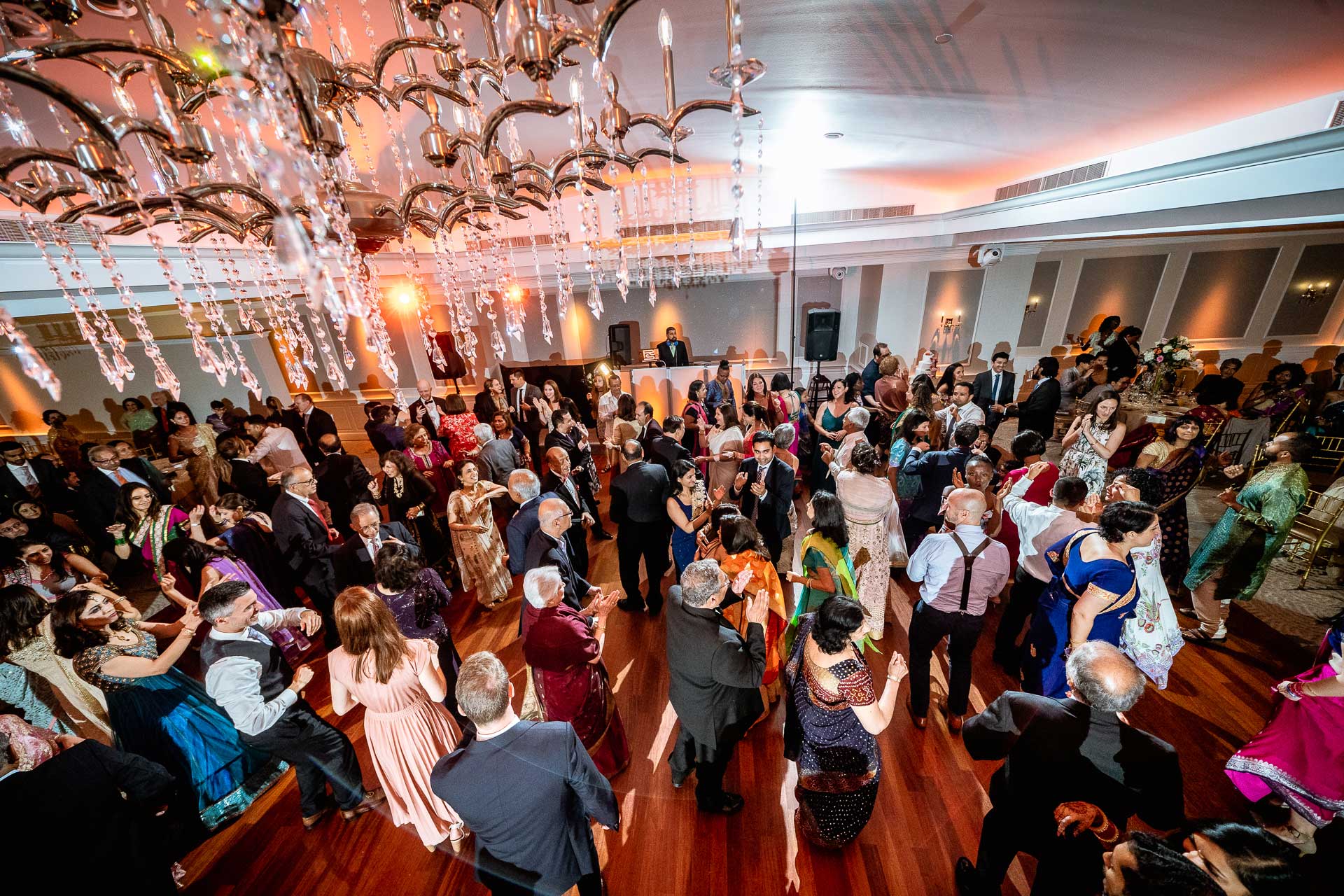 brooklake-Indian-Wedding-photographer-056 A large group of people in formal attire dance and mingle on a wooden floor under elaborate ceiling decorations at an indoor NJ Teen Events gathering.