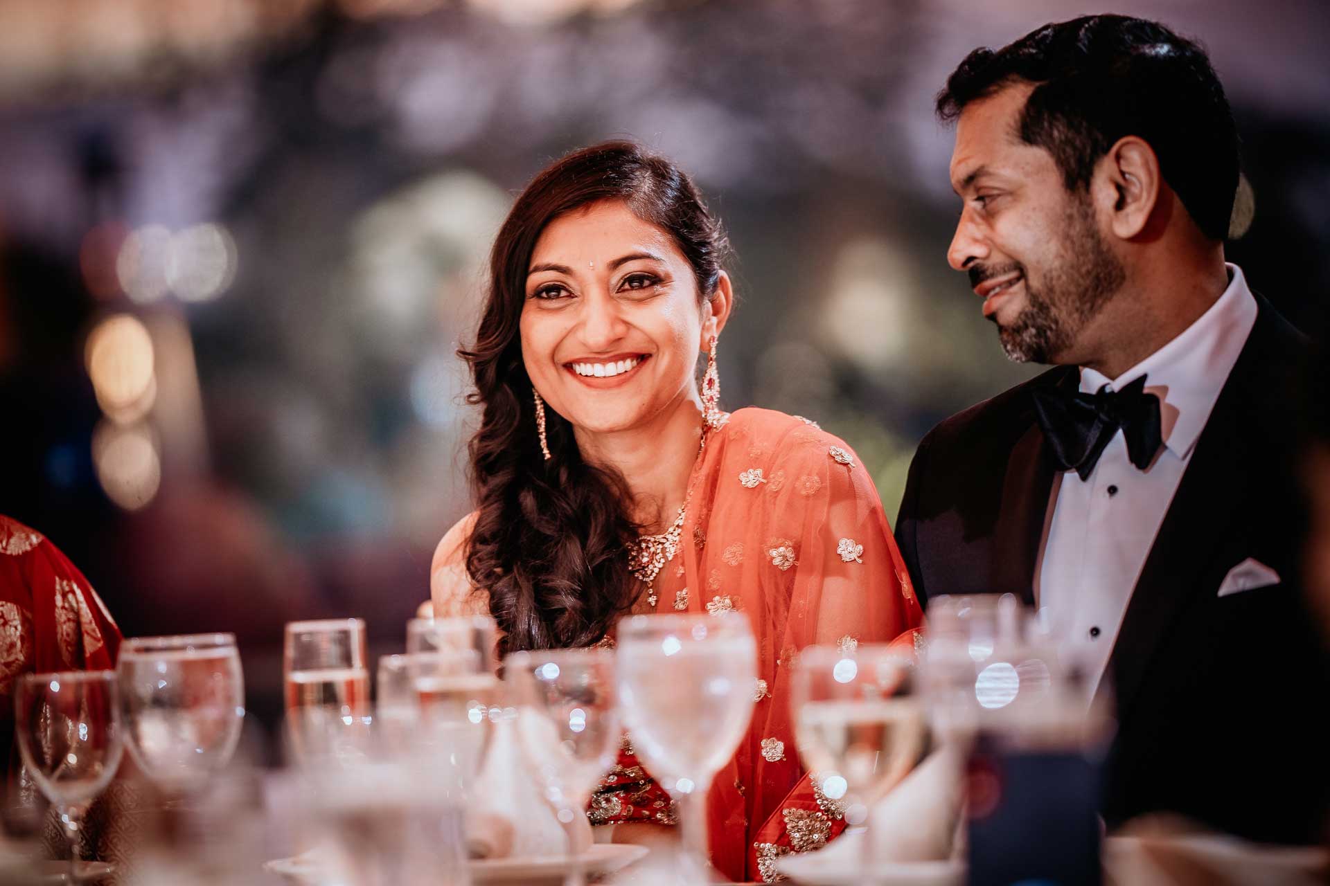 brooklake-Indian-Wedding-photographer-057 A woman in a red saree smiles at a formal NJ Teen Events gathering, seated beside a man in a black tuxedo. A dining table with glasses is in the foreground.